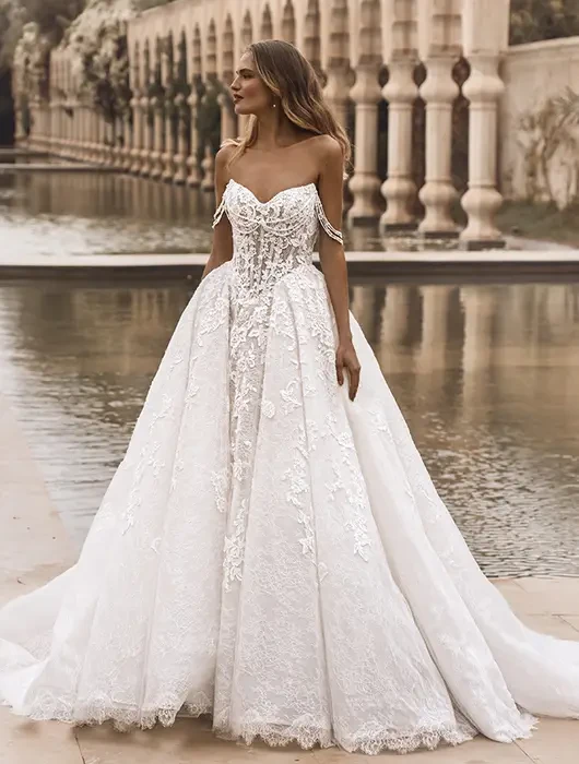 A model wearing an elegant white off-shoulder wedding gown with intricate lace details, standing near a decorative pool surrounded by columns and greenery.