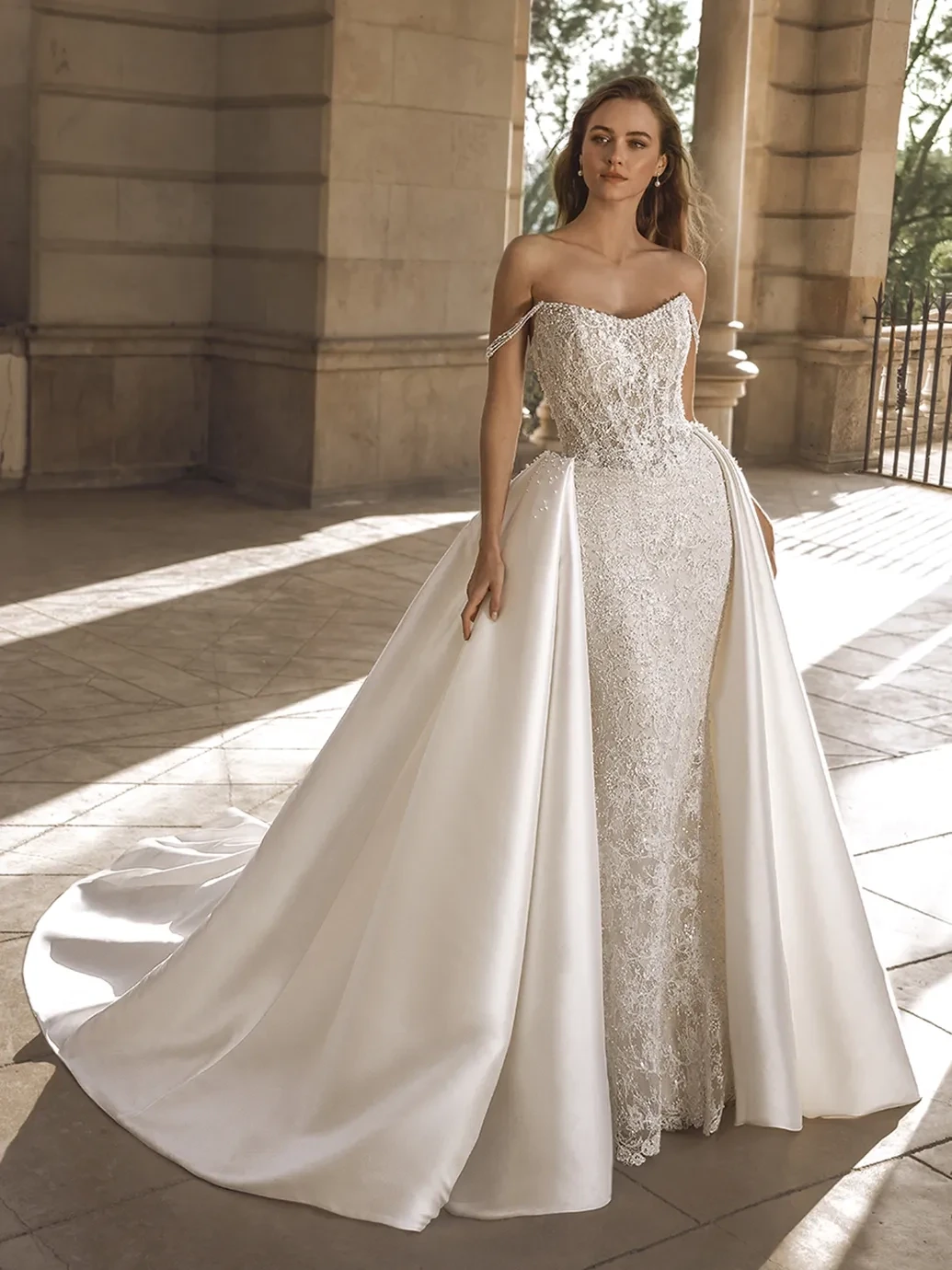 A model wears a strapless lace wedding gown with a satin overskirt in a sunlit stone archway.