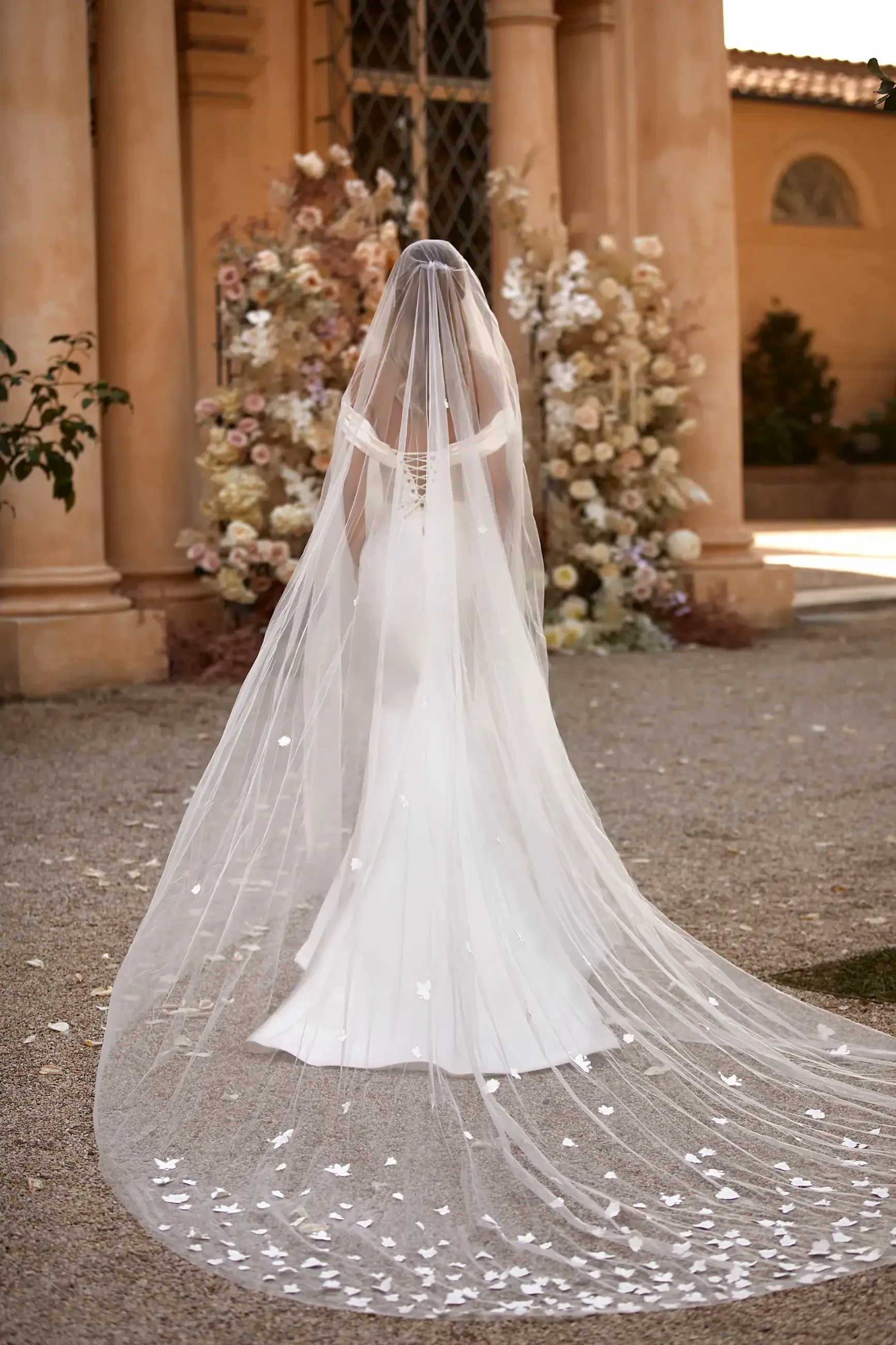 A bride in a white gown and long, flowing veil adorned with petals stands facing a floral backdrop, set against an ornate building, evoking elegance.
