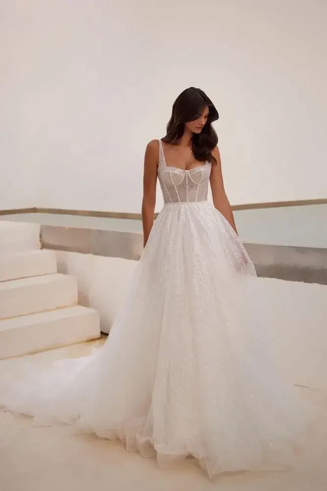 A woman in a shimmering white wedding gown stands gracefully on white steps. The dress features a fitted bodice and flowing skirt, exuding elegance and romance.