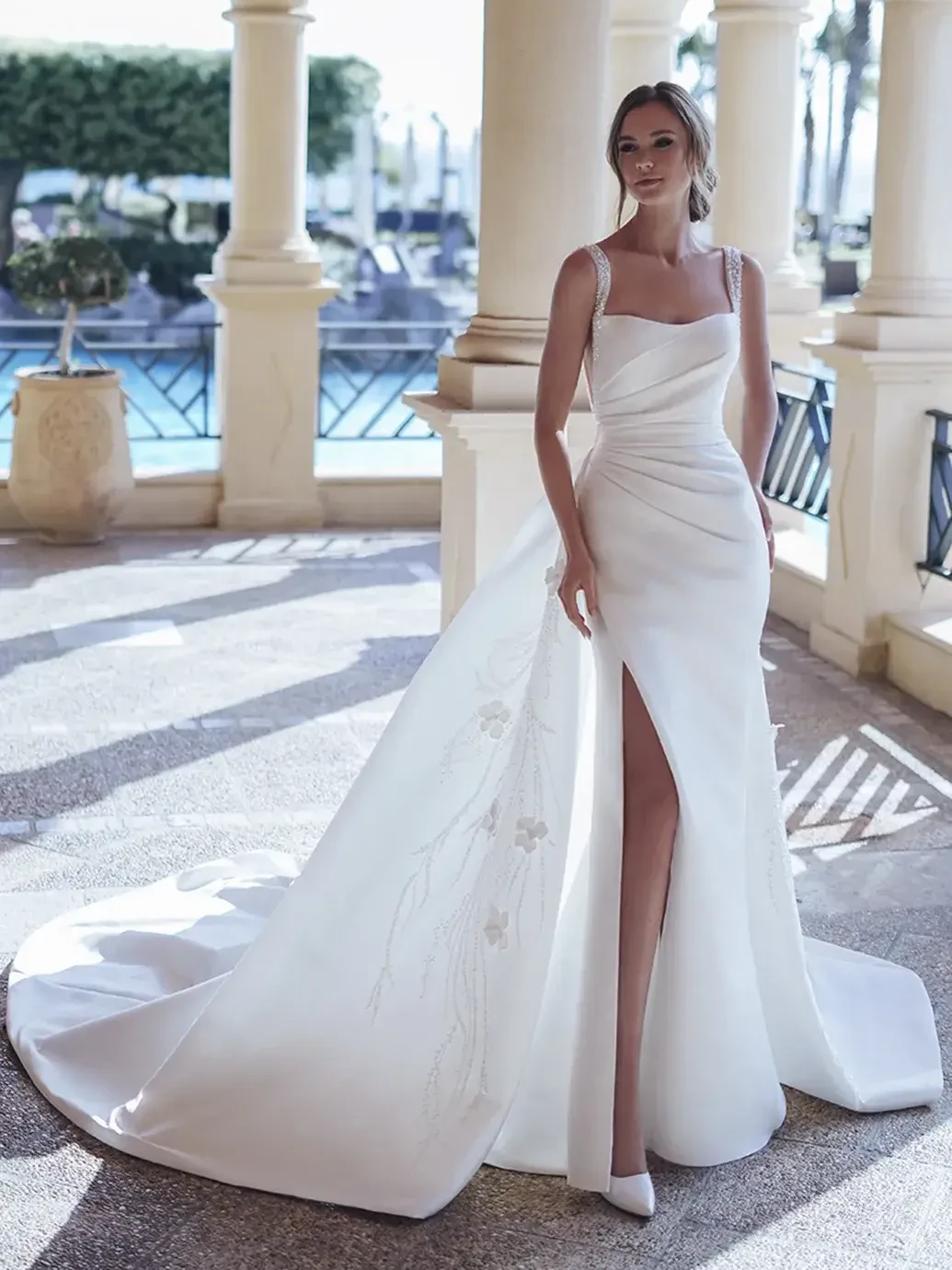 Bride in elegant white gown with a long train, standing on a sunlit terrace. The dress features a thigh-high slit and delicate floral embroidery, evoking a chic and sophisticated tone.