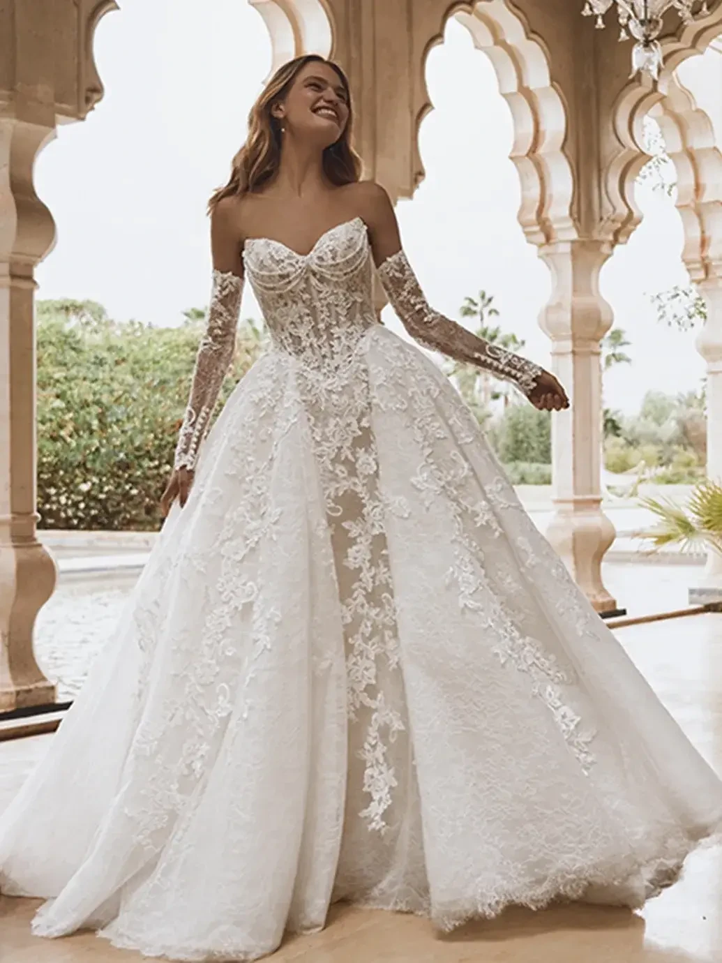 A woman in a beautiful white wedding gown with intricate lace detailing, modeled against an elegant architectural backdrop.